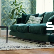 Un canapé vert avec des coussins fleuris dans un salon avec du papier peint vert et une plante luxuriante. Découvrez l'élégance parisienne avec une qualité inégalée, en ajoutant une touche de confort sophistiqué à votre vie à un prix exceptionnel. Une vie confortable. Canape Paris by Atelier Bis