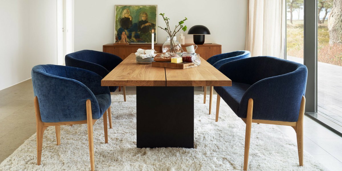 Une salle à manger chic avec une table en bois et quatre élégantes chaises en velours bleu, complétées par des œuvres d'art et une décoration sur un tapis blanc moelleux. De grandes fenêtres offrent une ambiance parisienne aérée, parfaite pour déguster des canapés entre amis. Canape Paris by Atelier Bis