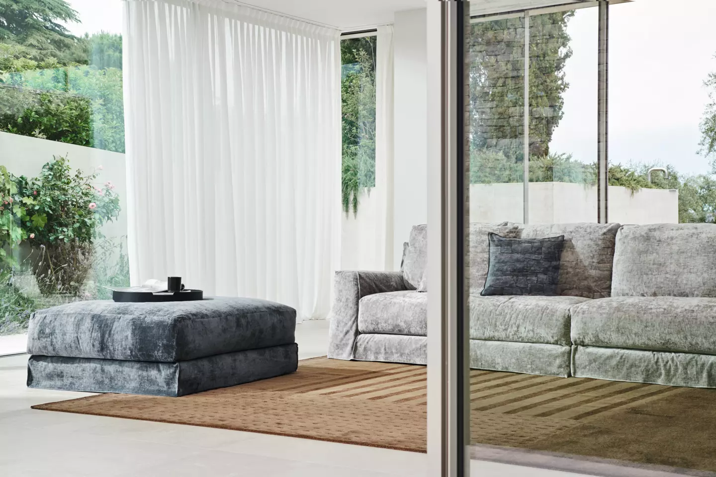 Salon moderne avec canapé gris, pouf assorti et tapis marron. De grandes fenêtres offrent une vue rappelant un jardin parisien, équilibrant parfaitement prix et qualité dans un cadre serein. Canape Paris by Atelier Bis