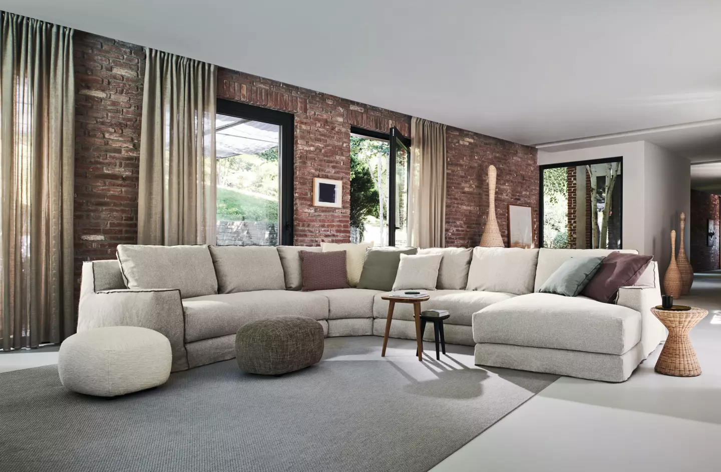 Découvrez un salon moderne qui respire la qualité d'une salle d'exposition, avec un grand canapé sectionnel beige contre des murs de briques rustiques et de grandes fenêtres ornées de rideaux élégants, apportant une touche de charme parisien dans votre espace. Canape Paris by Atelier Bis