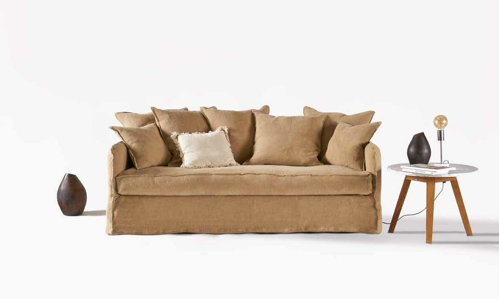 Canapé beige avec coussins, table d'appoint ronde sur laquelle repose une lampe et petit vase posé au sol. Décoration simple et moderne qui respire la qualité parisienne. Canape Paris by Atelier Bis