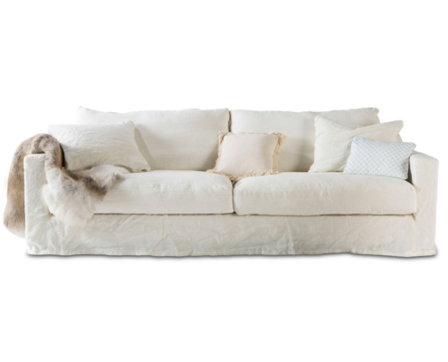 Un canapé blanc avec plusieurs coussins et un jeté de fourrure drapé sur un côté reflète l'élégance du showroom tout en alliant finesse et confort. Canape Paris by Atelier Bis