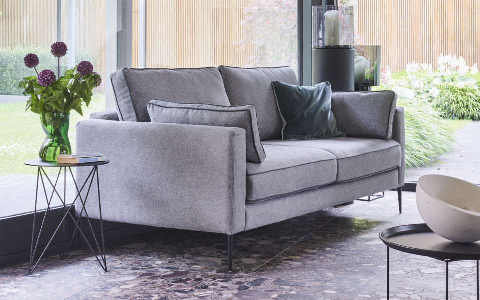 canape-4-places-peter-200-cm-3 Un canapé gris orne la salle d'exposition près des baies vitrées, accompagné d'une petite table d'appoint ornée d'un vase de fleurs et d'une pile de livres, mettant en valeur à la fois la qualité et l'élégance. Canape Paris by Atelier Bis