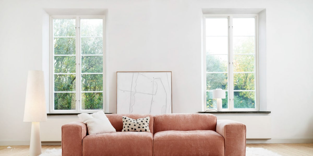 61cedc99d0a0a55b62ecb2c2_EDDA_interior_set1_wildflower_8_dust_pink_1 Un salon avec un canapé rose pâle, des coussins colorés et une table basse en métal doré.