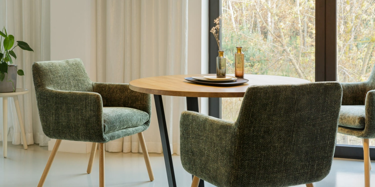 61ceddb3baf02f3f5da53e13_MOA_bistro_chair_bloom_6_green_3 La salle à manger comprend une table ronde en bois, des chaises rembourrées vertes et une plante en pot près de grandes fenêtres avec des rideaux, rappelant une salle d'exposition où chaque détail témoigne de l'équilibre parfait entre prix et qualité. Canape Paris by Atelier Bis