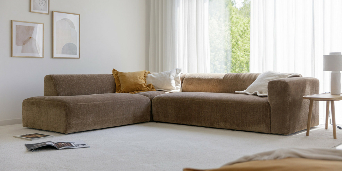 ANNIE_interior_chaiselongue_left_corner90_3seater_right_sky_4_gold_beige_1 Un salon cosy avec un canapé marron en L, des coussins, une table d'appoint et des œuvres d'art rappelant un showroom parisien, le tout baigné de lumière naturelle grâce à de grandes fenêtres. Canape Paris by Atelier Bis