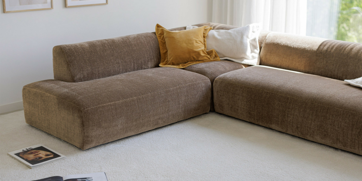 ANNIE_interior_chaiselongue_left_corner90_3seater_right_sky_4_gold_beige_2 Un canapé sectionnel marron avec des coussins trône dans une pièce lumineuse, faisant écho à la qualité que l'on retrouve dans un showroom haut de gamme. Des magazines sont éparpillés sur le sol recouvert de moquette, capturant le style pour toutes les gammes de prix. Canape Paris by Atelier Bis