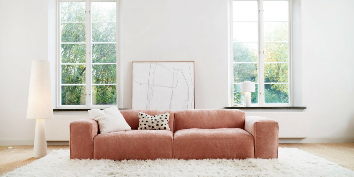 EDDA_interior_set1_wildflower_8_dust_pink_1 Une pièce avec un canapé en velours rose, des rideaux assortis et un tapis à motifs géométriques.