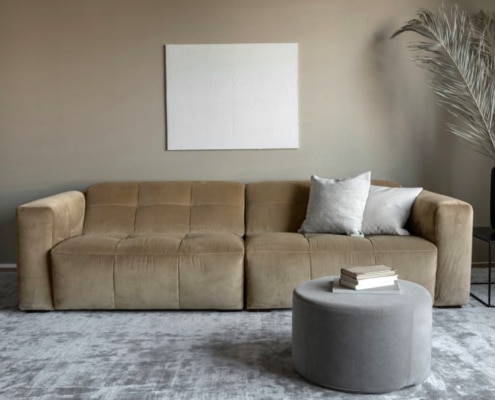 Un canapé beige avec des coussins blancs, une toile vierge rappelant l'élégance parisienne au-dessus, un pouf rond gris avec des livres et un vase avec de l'herbe de la pampa. Canape Paris by Atelier Bis