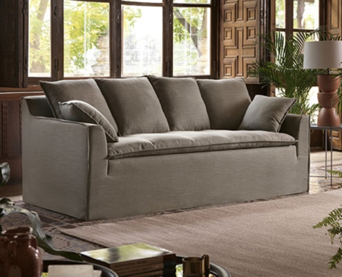 Un canapé gris avec des coussins trône élégamment dans une pièce cosy lambrissée de bois qui ressemble à un showroom parisien chic, agrémenté de grandes fenêtres et de plantes vibrantes. Canape Paris by Atelier Bis