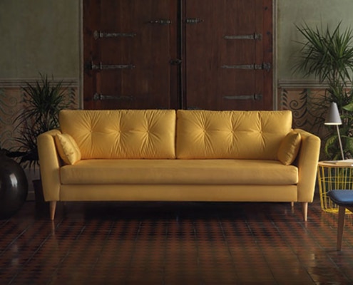 Un canapé jaune orne une pièce élégante ornée de plantes luxuriantes, de murs décoratifs et d'une porte en bois, dégageant un air de qualité indéniable. Canape Paris by Atelier Bis