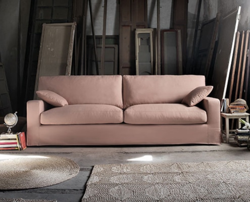 Un canapé beige orne la salle d'exposition, accompagné de deux coussins au milieu de livres, d'un globe et de divers cadres et portes en bois anciens, dégageant une qualité intemporelle. Canape Paris by Atelier Bis