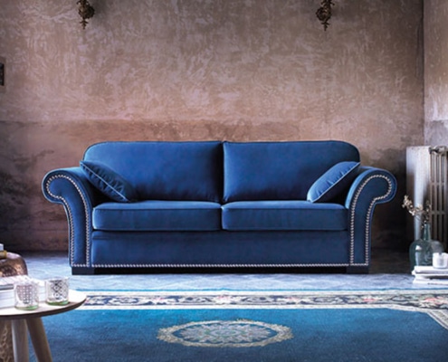 Un canapé en velours bleu avec coussins orne le salon rustique, rappelant l'élégance d'un showroom. Un tapis décoratif et une petite table complètent la scène, alliant harmonieusement prix et qualité. Canape Paris by Atelier Bis