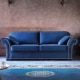 Un canapé en velours bleu avec coussins orne le salon rustique, rappelant l'élégance d'un showroom. Un tapis décoratif et une petite table complètent la scène, alliant harmonieusement prix et qualité. Canape Paris by Atelier Bis