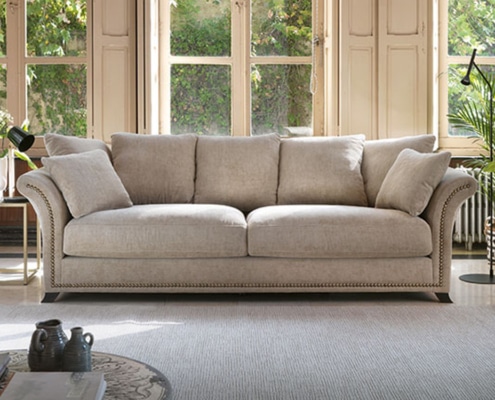 Un canapé beige avec des coussins trône élégamment dans un showroom parisien lumineux, près de grandes fenêtres et de plantes vertes vibrantes, mettant en valeur son savoir-faire de qualité. Canape Paris by Atelier Bis