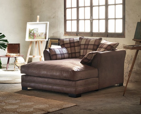 Une chaise longue en cuir marron avec des coussins à carreaux trône de manière accueillante dans une pièce rustique, où de grandes fenêtres et un chevalet en bois rehaussent l'ambiance de qualité showroom. Canape Paris by Atelier Bis