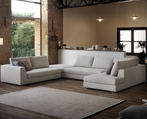 Un salon moderne avec un grand canapé beige, un parquet en bois et un tapis élégant, le tout baigné d'un éclairage chaleureux près d'une grande fenêtre. Son décor respire l'élégance et la qualité parisiennes, créant un mélange parfait de confort et de style. Canape Paris by Atelier Bis