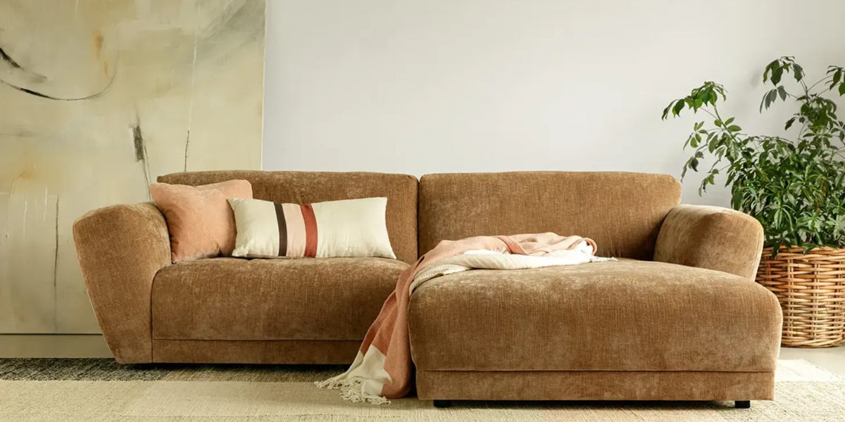 ASTA_SITS_canape-paris_02 Une photo du canapé Asta marron dans un salon de style industriel avec des murs en briques apparentes et des étagères en métal pour une ambiance urbaine et moderne.