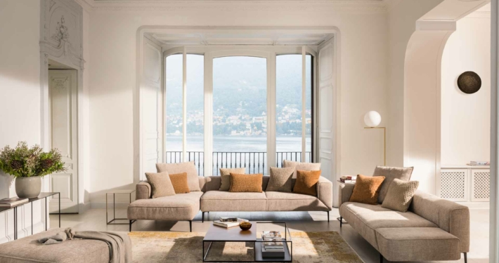 Un salon spacieux avec des canapés beiges de haute qualité et des coussins moelleux autour d'une table basse élégante. De grandes fenêtres baignent l'espace de lumière naturelle, offrant une vue imprenable sur le lac et les montagnes - une touche d'élégance parisienne dans chaque coin. Canape Paris by Atelier Bis