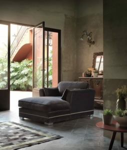 Une pièce cosy et tamisée à Paris présente un grand canapé sombre et des plantes en pot, avec une porte ouverte donnant sur un jardin luxuriant. La scène incarne à la fois la qualité et le charme. Canape Paris by Atelier Bis