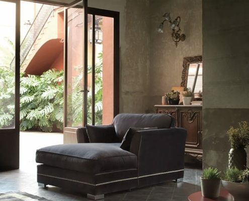 Une pièce cosy et tamisée à Paris présente un grand canapé sombre et des plantes en pot, avec une porte ouverte donnant sur un jardin luxuriant. La scène incarne à la fois la qualité et le charme. Canape Paris by Atelier Bis