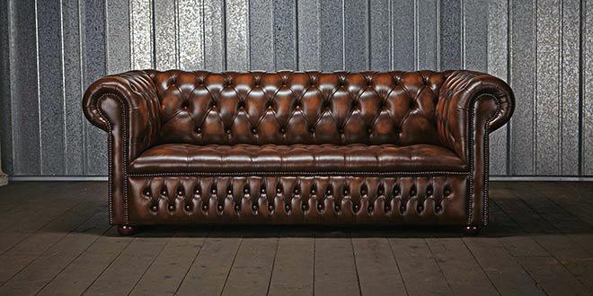 Un canapé Chesterfield en cuir marron repose élégamment contre un mur en métal ondulé sur un plancher en bois, dégageant un air de qualité parisienne. Canape Paris by Atelier Bis